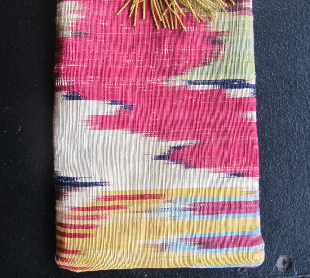 A rectangular double-sided silk ikat purse featuring colourful horizontal bands in pink, yellow, blue, and cream with a red tassel attached at the top opening. View VI