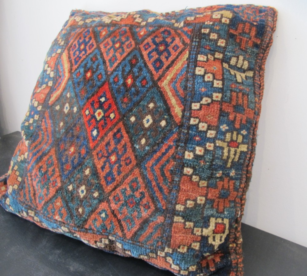 A square handwoven Kurdish wool cushion featuring geometric diamond motifs in blue, terracotta, and brown shades.