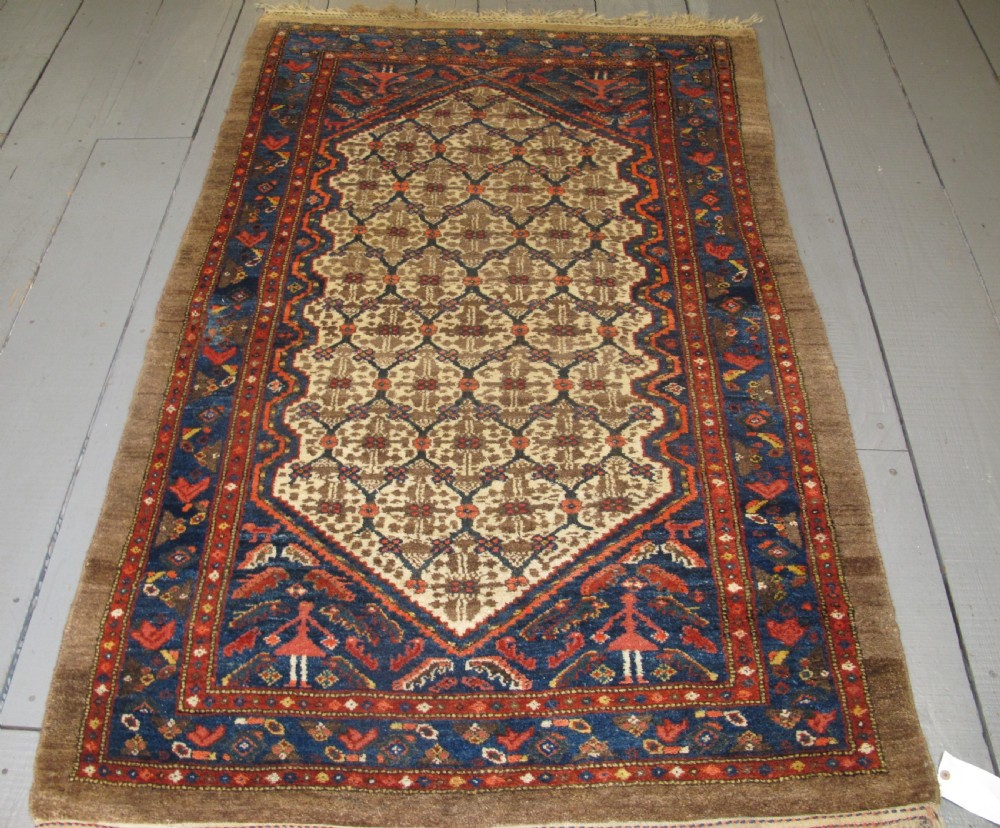 An antique Persian Kolyai hand-knotted wool rug displaying a cream lattice central medallion framed by a deep blue field with stylized figures and tan borders.