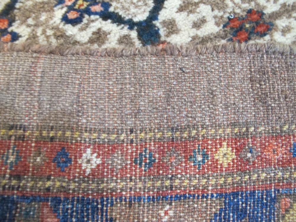 An antique Persian Kolyai hand-knotted wool rug displaying a cream lattice central medallion framed by a deep blue field with stylized figures and tan borders. View X