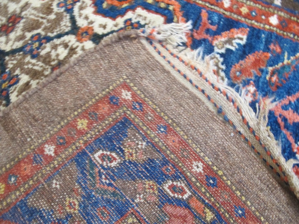 An antique Persian Kolyai hand-knotted wool rug displaying a cream lattice central medallion framed by a deep blue field with stylized figures and tan borders. View XI