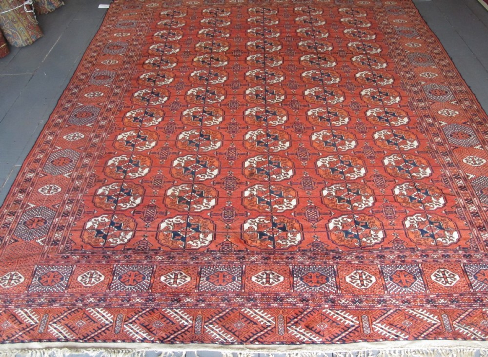 A large hand-knotted antique wool carpet featuring repeating octagonal Tekke gul motifs in rust red, cream, and navy blue with geometric borders.