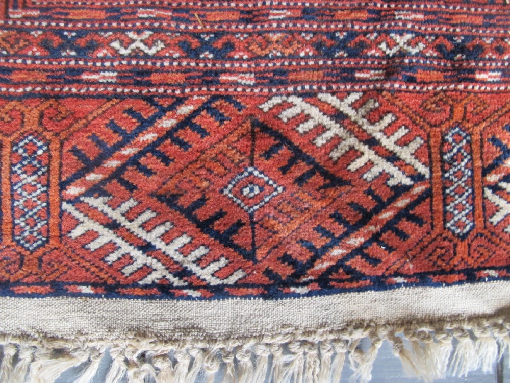 A large hand-knotted antique wool carpet featuring repeating octagonal Tekke gul motifs in rust red, cream, and navy blue with geometric borders. View IV