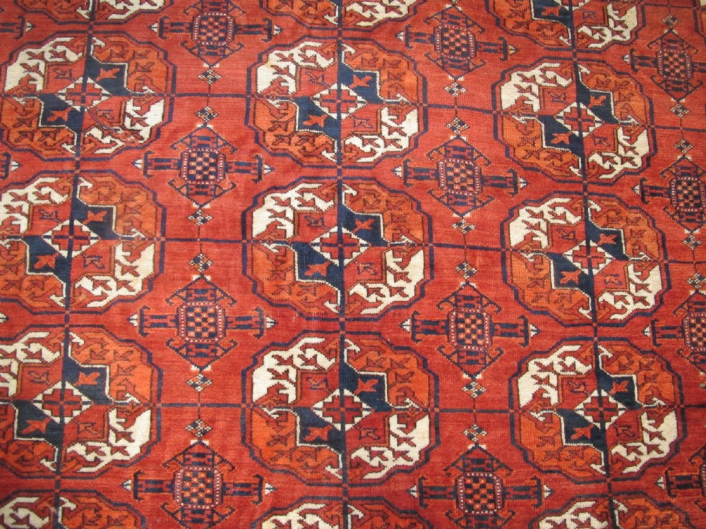 A large hand-knotted antique wool carpet featuring repeating octagonal Tekke gul motifs in rust red, cream, and navy blue with geometric borders. View V