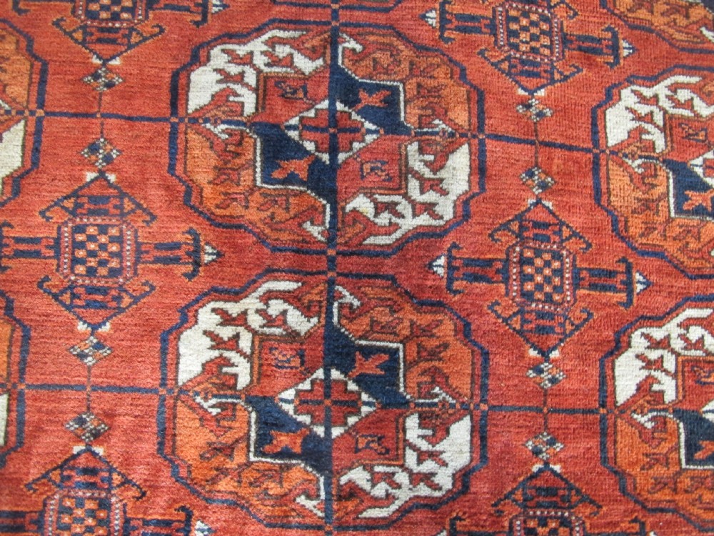 A large hand-knotted antique wool carpet featuring repeating octagonal Tekke gul motifs in rust red, cream, and navy blue with geometric borders. View VI