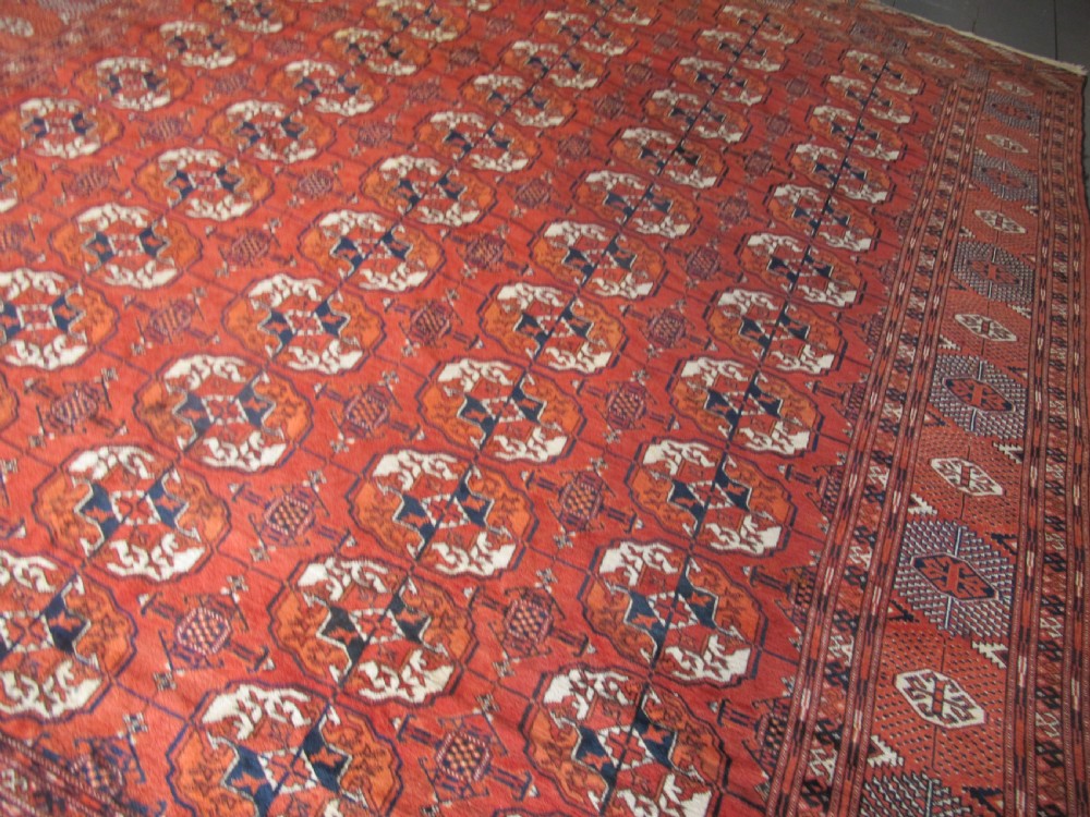 A large hand-knotted antique wool carpet featuring repeating octagonal Tekke gul motifs in rust red, cream, and navy blue with geometric borders. View VII