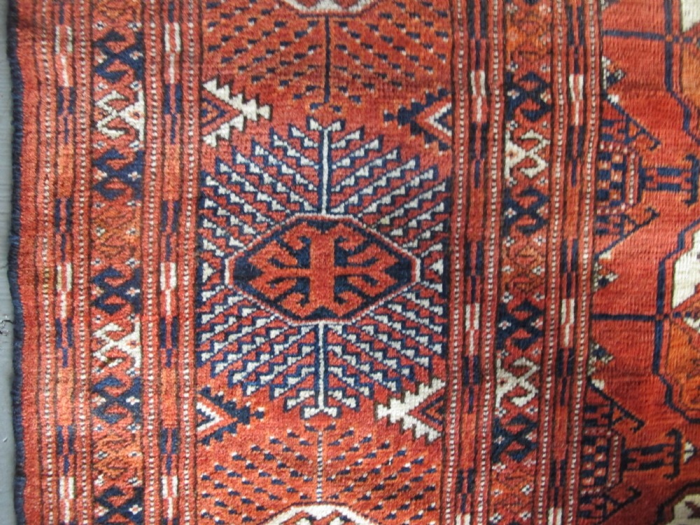 A large hand-knotted antique wool carpet featuring repeating octagonal Tekke gul motifs in rust red, cream, and navy blue with geometric borders. View VIII