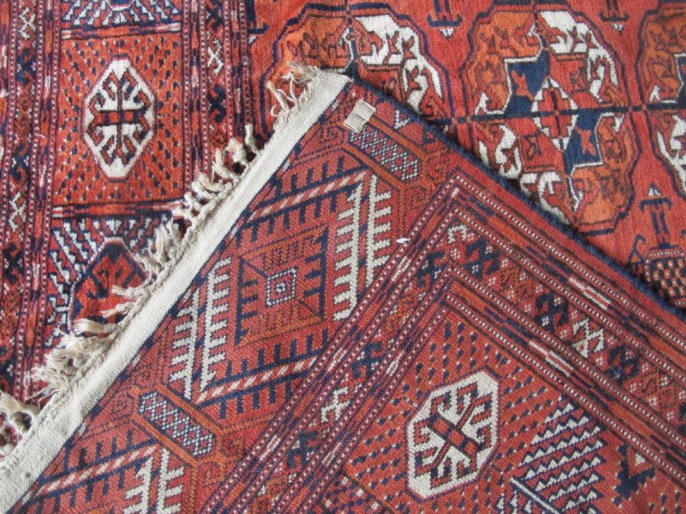 A large hand-knotted antique wool carpet featuring repeating octagonal Tekke gul motifs in rust red, cream, and navy blue with geometric borders. View X