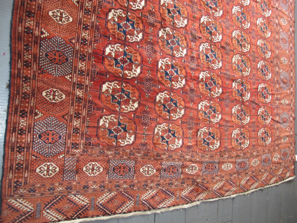 A large hand-knotted antique wool carpet featuring repeating octagonal Tekke gul motifs in rust red, cream, and navy blue with geometric borders. View XII