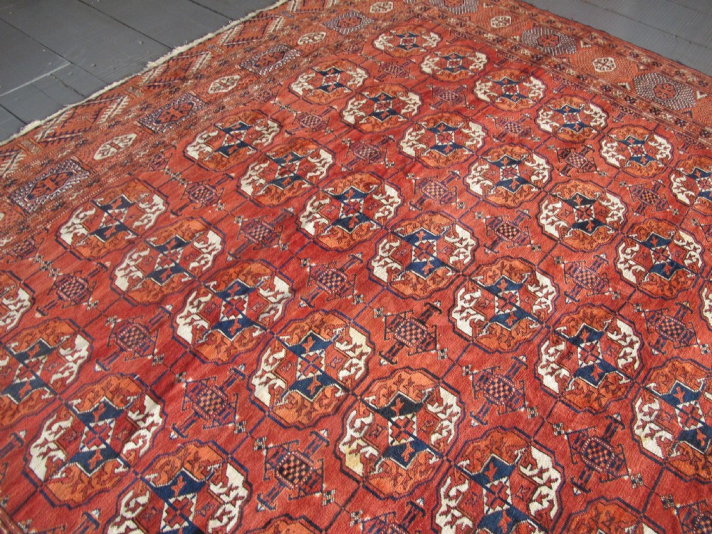 A large hand-knotted antique wool carpet featuring repeating octagonal Tekke gul motifs in rust red, cream, and navy blue with geometric borders. View XV