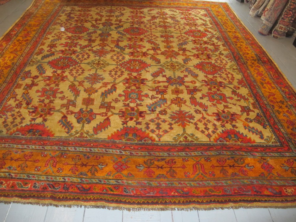 A large hand-knotted wool Oushak carpet featuring a golden-yellow field with geometric medallion motifs bordered by wide terracotta and orange patterned bands.