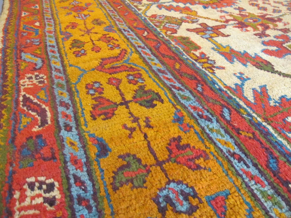 A large hand-knotted wool Oushak carpet featuring a golden-yellow field with geometric medallion motifs bordered by wide terracotta and orange patterned bands. View VI