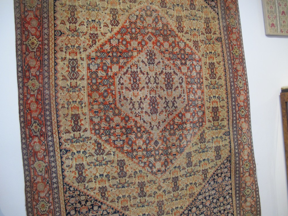 An antique Persian Senneh wool carpet featuring a large central diamond medallion surrounded by fine geometric patterning in terracotta, navy, and cream tones. View X