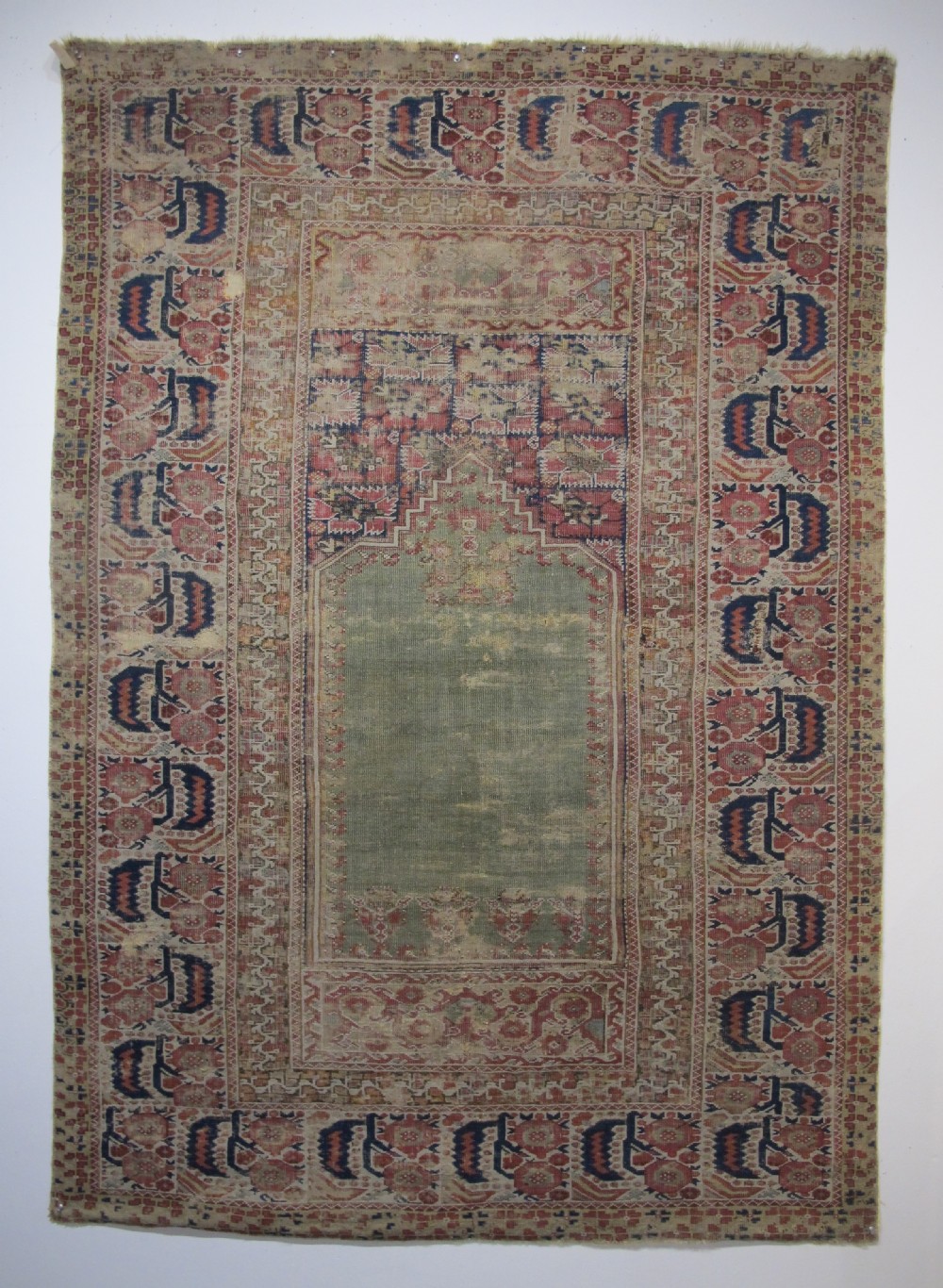 An 18th-century hand-knotted Ghiordes prayer rug featuring a central pale green mihrab niche framed by intricate floral borders in red, blue, and beige.