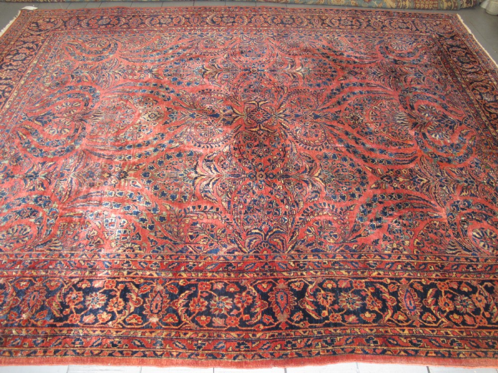 A large hand-knotted Persian Sarouk carpet featuring a dense floral pattern on a rose red field with a navy blue border.