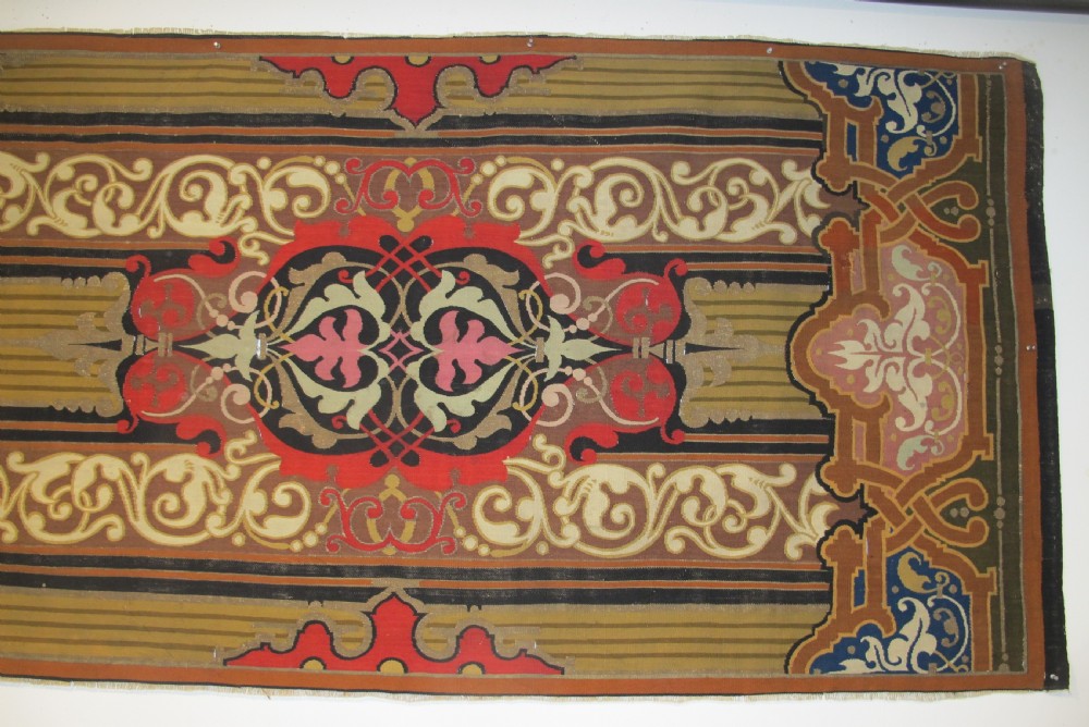 A long horizontal European wool tapestry panel featuring ornate scrolling foliage, geometric motifs, and metallic thread detailing in tones of red, olive, and beige. View IV