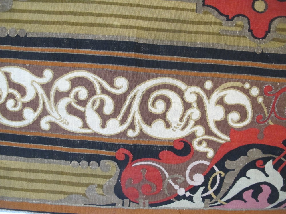 A long horizontal European wool tapestry panel featuring ornate scrolling foliage, geometric motifs, and metallic thread detailing in tones of red, olive, and beige. View VII