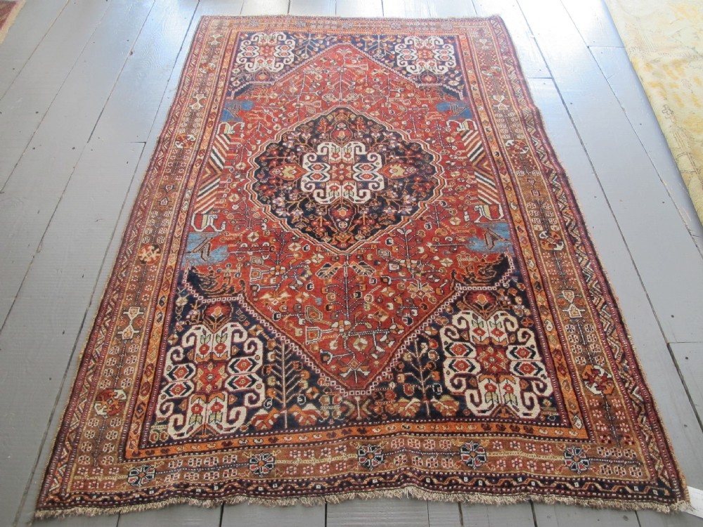 An antique Qashqai wool rug featuring a central diamond medallion, geometric tribal motifs, and intricate borders in shades of terracotta red, navy indigo, and cream.