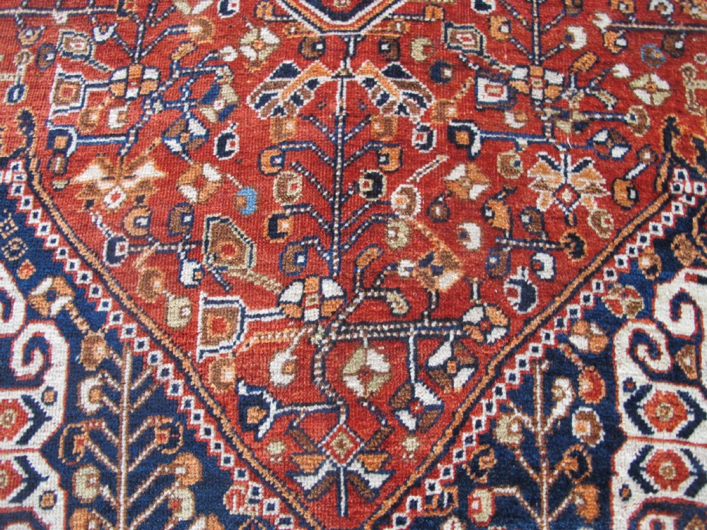 An antique Qashqai wool rug featuring a central diamond medallion, geometric tribal motifs, and intricate borders in shades of terracotta red, navy indigo, and cream. View V