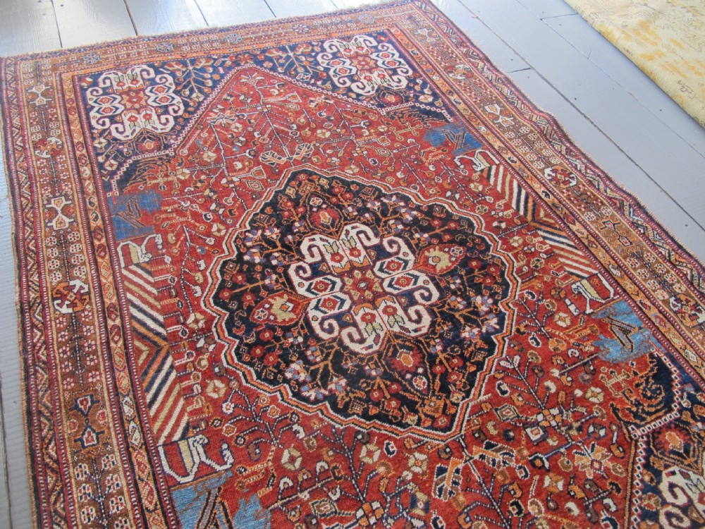 An antique Qashqai wool rug featuring a central diamond medallion, geometric tribal motifs, and intricate borders in shades of terracotta red, navy indigo, and cream. View X