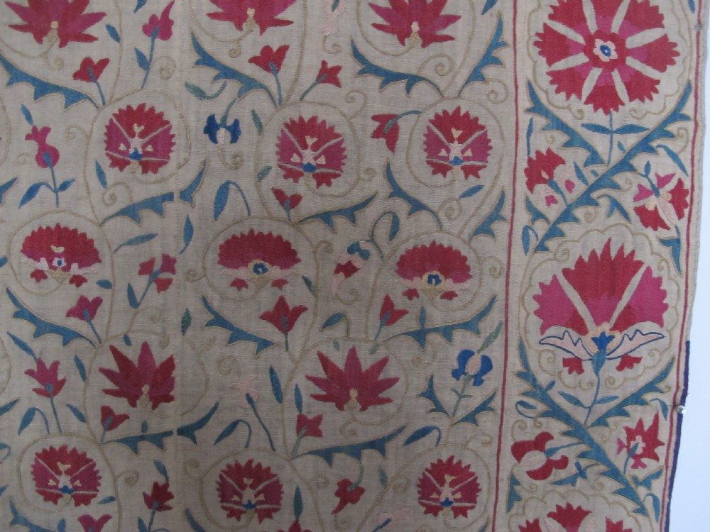 A rectangular antique silk-embroidered Suzani textile featuring crimson floral motifs and scrolling leaf borders on a beige cotton ground. View V