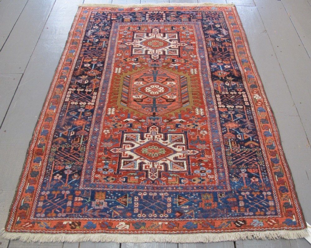 A rectangular hand-knotted antique Persian Karaja rug featuring three geometric medallions on a terracotta red field framed by a wide navy blue border with tribal motifs.