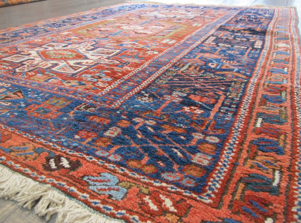 A rectangular hand-knotted antique Persian Karaja rug featuring three geometric medallions on a terracotta red field framed by a wide navy blue border with tribal motifs. View II