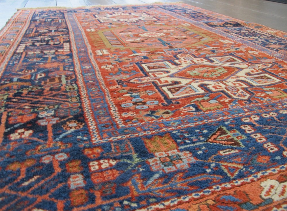 A rectangular hand-knotted antique Persian Karaja rug featuring three geometric medallions on a terracotta red field framed by a wide navy blue border with tribal motifs. View III