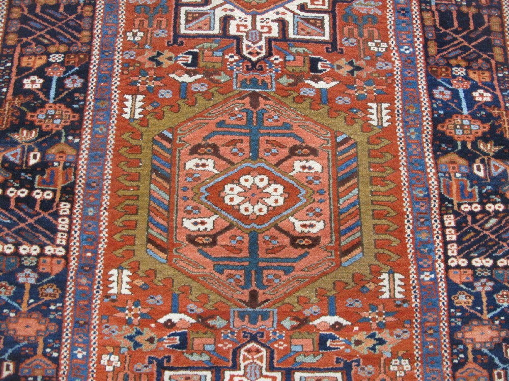 A rectangular hand-knotted antique Persian Karaja rug featuring three geometric medallions on a terracotta red field framed by a wide navy blue border with tribal motifs. View IV