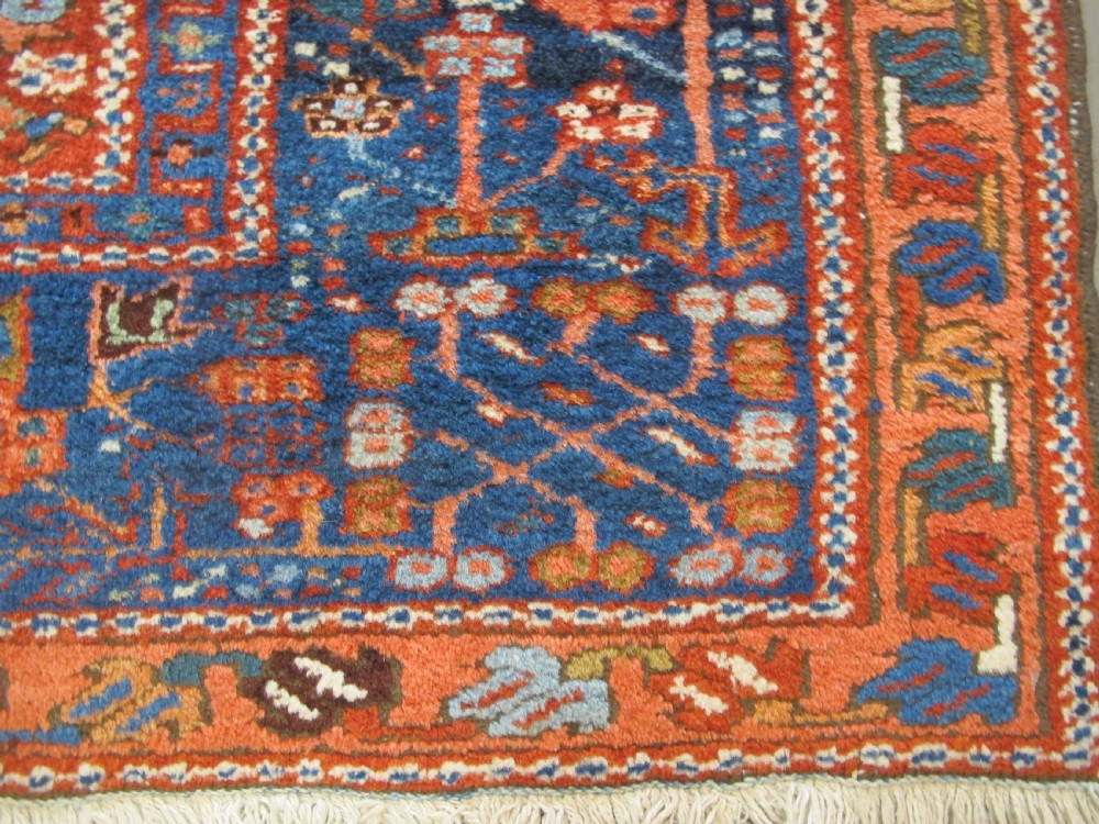 A rectangular hand-knotted antique Persian Karaja rug featuring three geometric medallions on a terracotta red field framed by a wide navy blue border with tribal motifs. View V