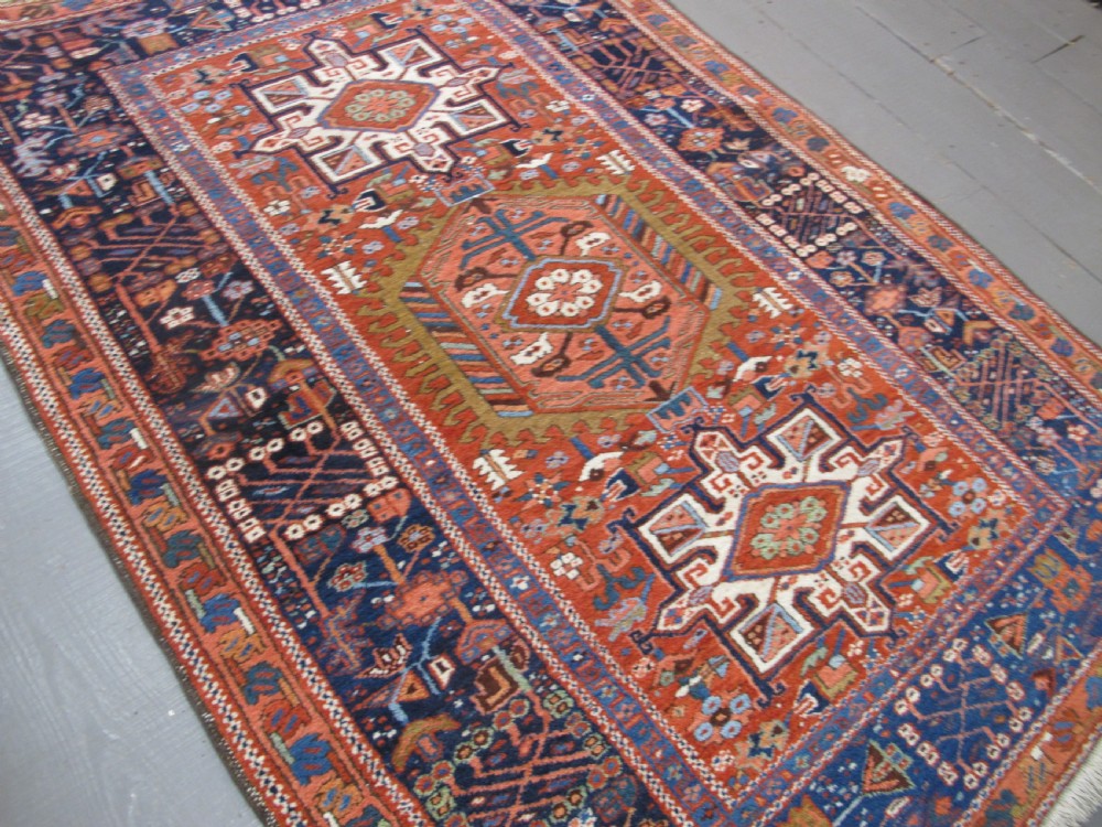 A rectangular hand-knotted antique Persian Karaja rug featuring three geometric medallions on a terracotta red field framed by a wide navy blue border with tribal motifs. View VI