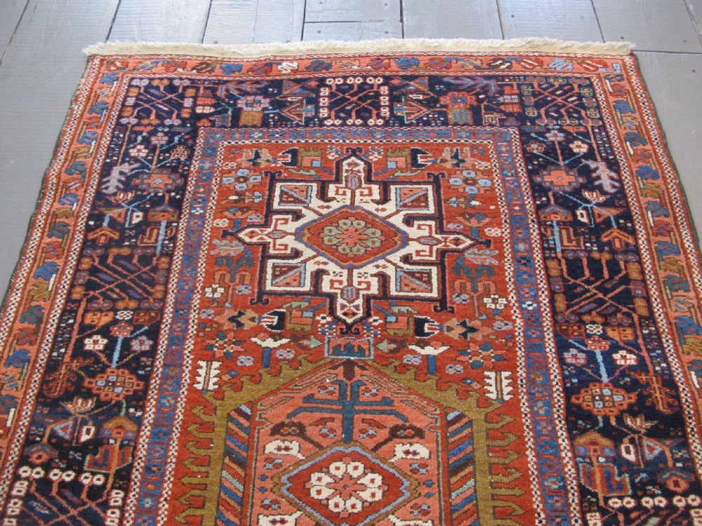 A rectangular hand-knotted antique Persian Karaja rug featuring three geometric medallions on a terracotta red field framed by a wide navy blue border with tribal motifs. View VII