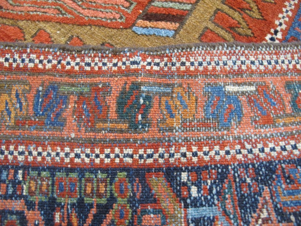 A rectangular hand-knotted antique Persian Karaja rug featuring three geometric medallions on a terracotta red field framed by a wide navy blue border with tribal motifs. View VIII