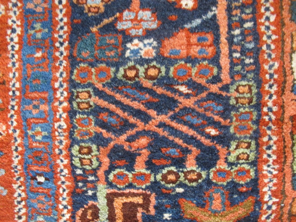 A rectangular hand-knotted antique Persian Karaja rug featuring three geometric medallions on a terracotta red field framed by a wide navy blue border with tribal motifs. View IX