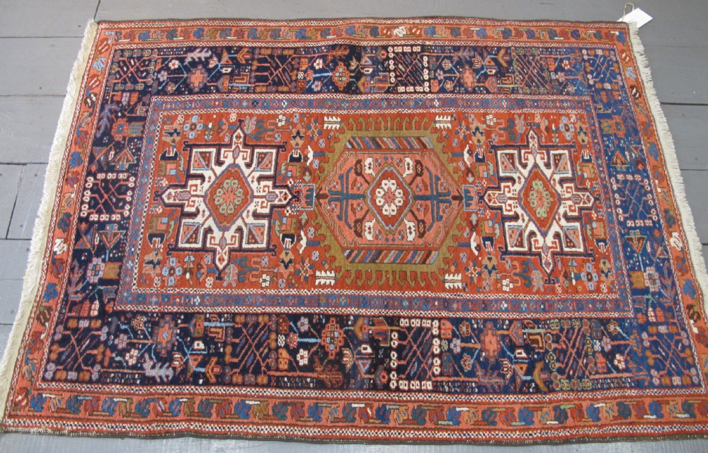 A rectangular hand-knotted antique Persian Karaja rug featuring three geometric medallions on a terracotta red field framed by a wide navy blue border with tribal motifs. View X