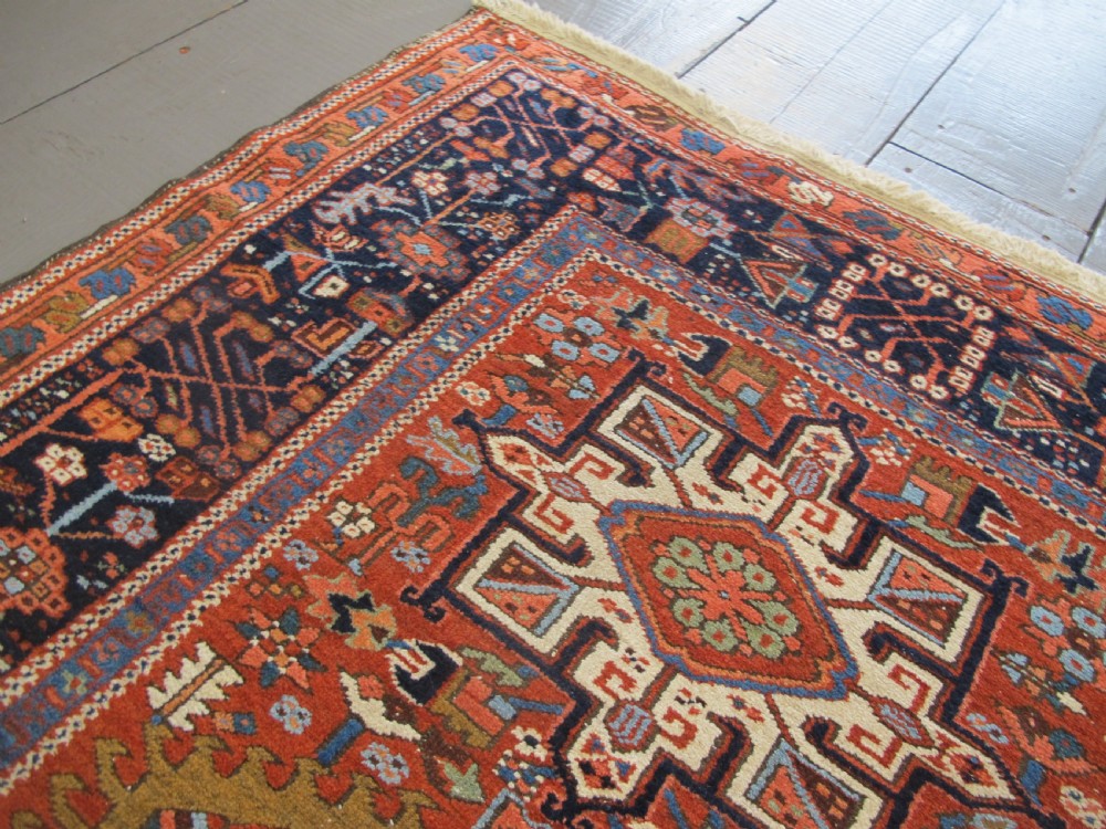 A rectangular hand-knotted antique Persian Karaja rug featuring three geometric medallions on a terracotta red field framed by a wide navy blue border with tribal motifs. View XI