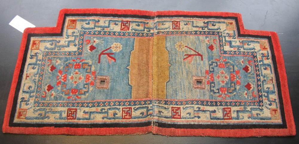 A hand-knotted wool Tibetan saddle cover featuring a blue central field with floral motifs, geometric inner borders, a red outer margin, and leather-bound stirrup slots.