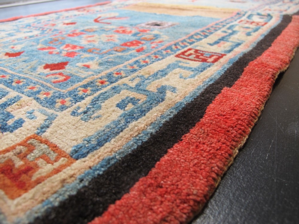 A hand-knotted wool Tibetan saddle cover featuring a blue central field with floral motifs, geometric inner borders, a red outer margin, and leather-bound stirrup slots. View II