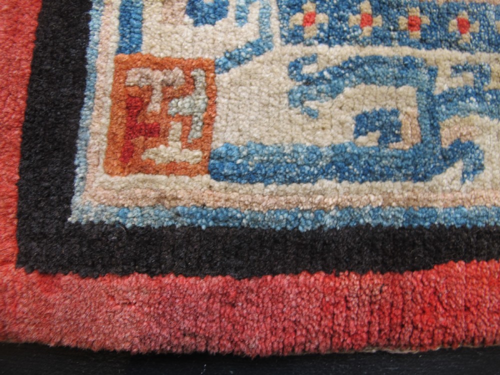 A hand-knotted wool Tibetan saddle cover featuring a blue central field with floral motifs, geometric inner borders, a red outer margin, and leather-bound stirrup slots. View III