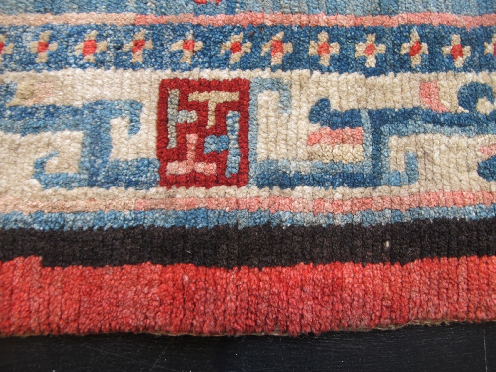 A hand-knotted wool Tibetan saddle cover featuring a blue central field with floral motifs, geometric inner borders, a red outer margin, and leather-bound stirrup slots. View IV