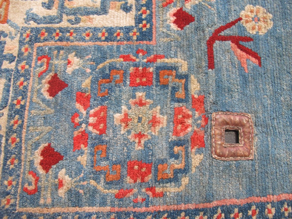 A hand-knotted wool Tibetan saddle cover featuring a blue central field with floral motifs, geometric inner borders, a red outer margin, and leather-bound stirrup slots. View V