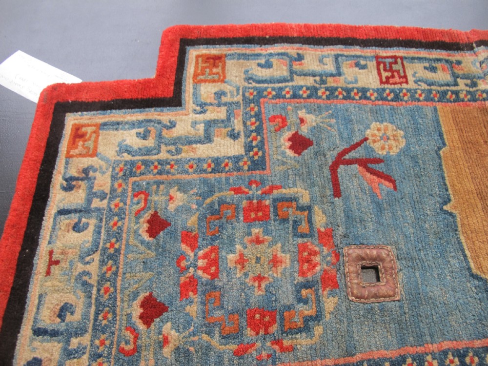 A hand-knotted wool Tibetan saddle cover featuring a blue central field with floral motifs, geometric inner borders, a red outer margin, and leather-bound stirrup slots. View VI