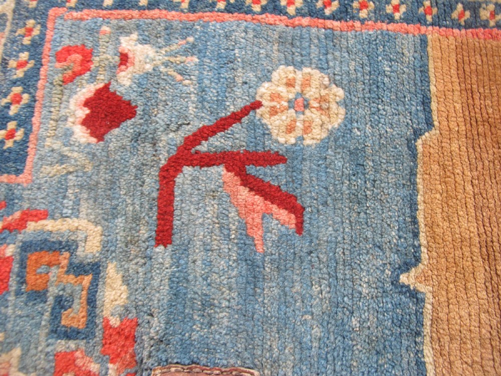 A hand-knotted wool Tibetan saddle cover featuring a blue central field with floral motifs, geometric inner borders, a red outer margin, and leather-bound stirrup slots. View VII
