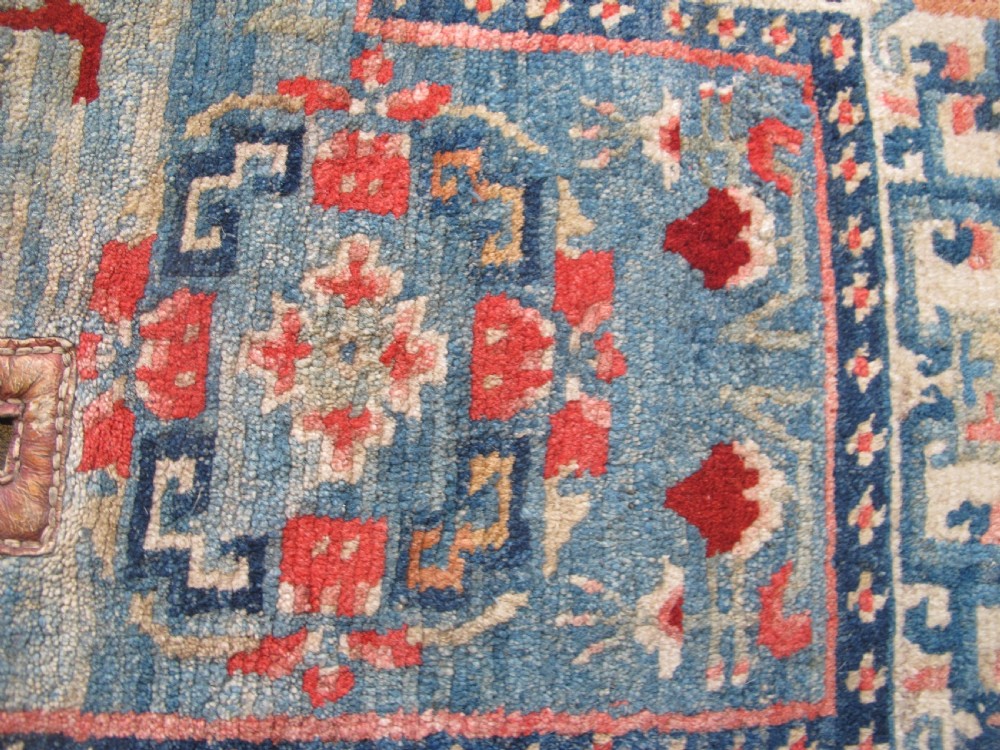 A hand-knotted wool Tibetan saddle cover featuring a blue central field with floral motifs, geometric inner borders, a red outer margin, and leather-bound stirrup slots. View IX
