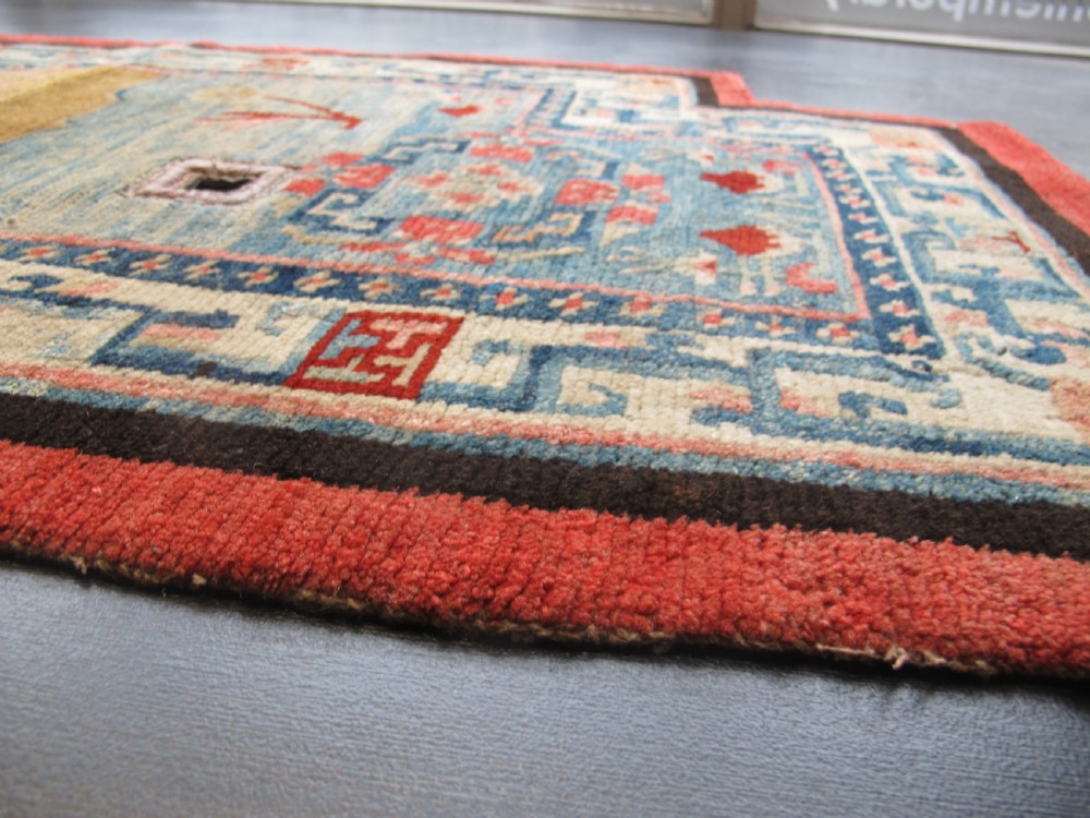 A hand-knotted wool Tibetan saddle cover featuring a blue central field with floral motifs, geometric inner borders, a red outer margin, and leather-bound stirrup slots. View X