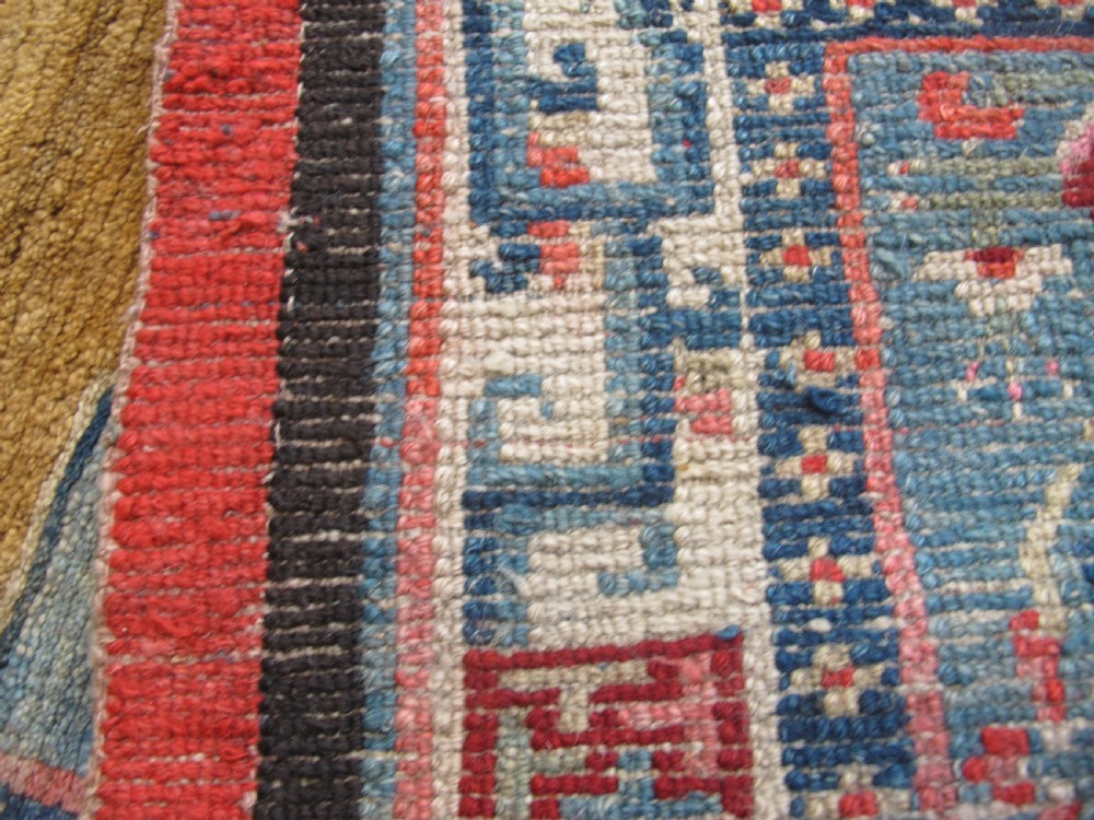 A hand-knotted wool Tibetan saddle cover featuring a blue central field with floral motifs, geometric inner borders, a red outer margin, and leather-bound stirrup slots. View XI