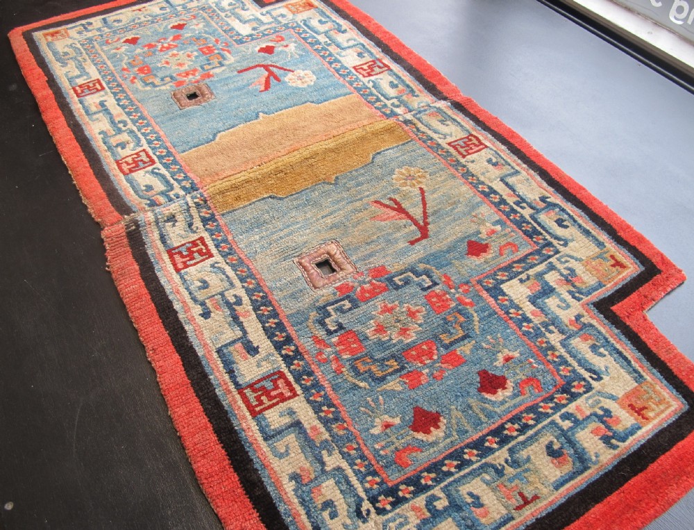 A hand-knotted wool Tibetan saddle cover featuring a blue central field with floral motifs, geometric inner borders, a red outer margin, and leather-bound stirrup slots. View XII