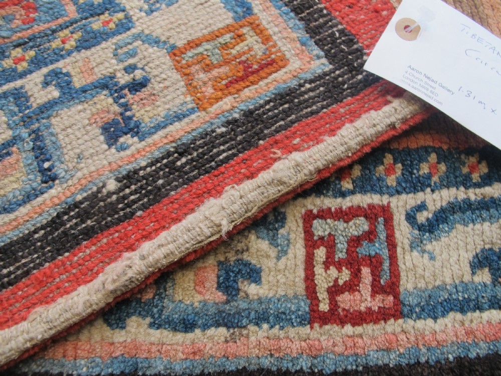 A hand-knotted wool Tibetan saddle cover featuring a blue central field with floral motifs, geometric inner borders, a red outer margin, and leather-bound stirrup slots. View XIII