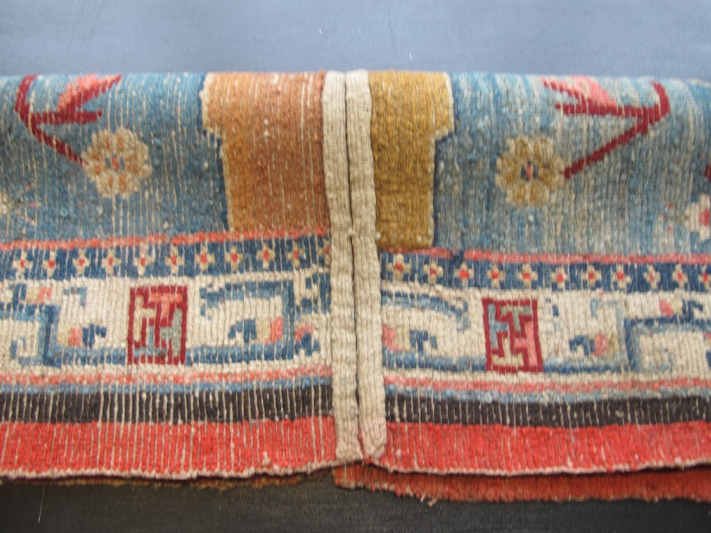 A hand-knotted wool Tibetan saddle cover featuring a blue central field with floral motifs, geometric inner borders, a red outer margin, and leather-bound stirrup slots. View XIV