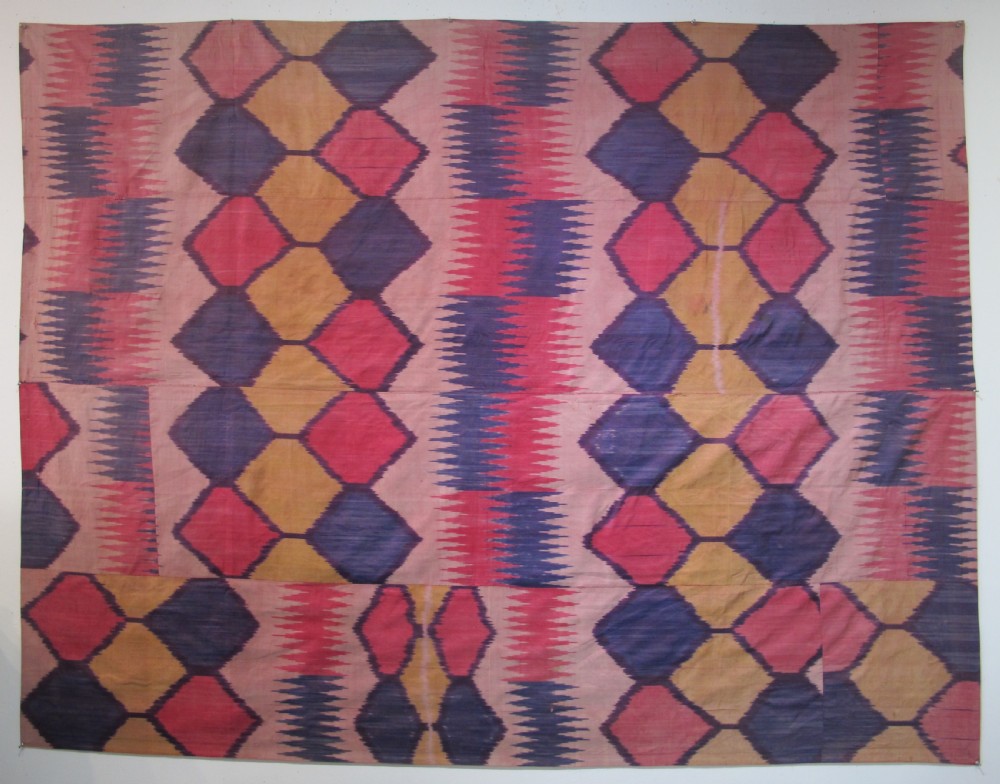 A large handwoven silk ikat textile panel from Uzbekistan featuring geometric diamond motifs and sawtooth patterns in pink, yellow, and purple.